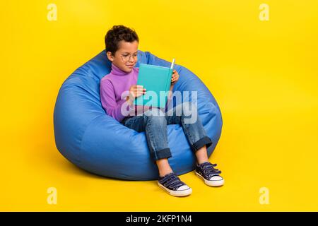 Grand portrait d'un écolier élégant assis sac confortable lire livre isolé sur fond jaune Banque D'Images