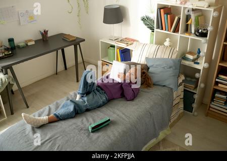 Vue en grand angle d'une petite fille qui s'est allongée sur son lit et qui lit son journal pendant son temps libre à la maison Banque D'Images