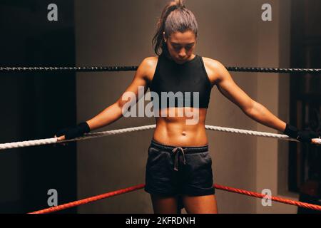 Boxeur féminin pensif debout dans un coin d'un anneau de boxe sans gants. Jeune femme sportive ayant une séance d'entraînement dans un gymnase de boxe. Banque D'Images