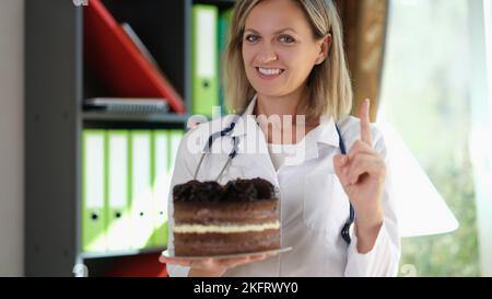 La femme médecin tient le gâteau au chocolat dans sa main et rappelle de ne pas manger de la nourriture indésirable. Banque D'Images