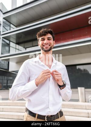 Un homme barbu souriant se tenant près d'un immeuble urbain à plusieurs étages et se tenant en contrebas d'une chemise tout en regardant l'appareil photo Banque D'Images