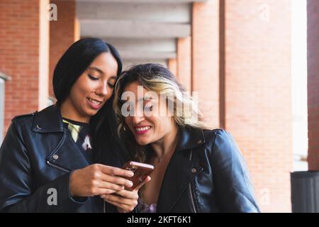 Heureuse femme hispanique dans une veste en cuir souriant et partageant des informations dans les médias sociaux avec une petite amie tout en se tenant à l'extérieur de la brique de construire ensemble Banque D'Images