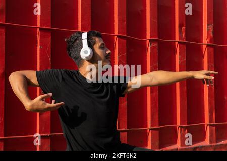 Homme énergique en costume noir dansant tout en écoutant la chanson préférée sur le casque contre la clôture en métal rouge Banque D'Images