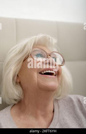 Bonne femme âgée en lunettes et chandail assis sur un lit confortable et doux Banque D'Images