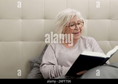 Bonne vieille dame en verres et chaud chandail assis sur le lit parmi les oreillers et lecture drôle de livre Banque D'Images