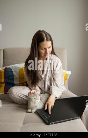 Femme en pyjama caressant Scottish Fold Cat et en parcourant un netbook tout en étant assis sur un canapé et en travaillant à distance dans le salon à la maison Banque D'Images