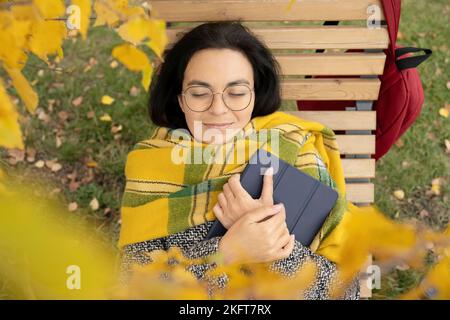 Grand angle de jeune femme dans des vêtements décontractés et lunettes de vue avec tablette numérique se détendre sur un lit de soleil en bois dans le parc Banque D'Images