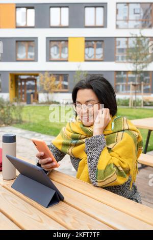 Jeune femme utilisant des écouteurs et un téléphone portable assis à table avec une tablette et un en-cas pour déjeuner dans le parc du campus en automne Banque D'Images