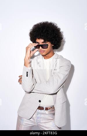 Jeune homme hispanique en costume élégant et lunettes de soleil regardant l'appareil photo debout sur fond gris Banque D'Images