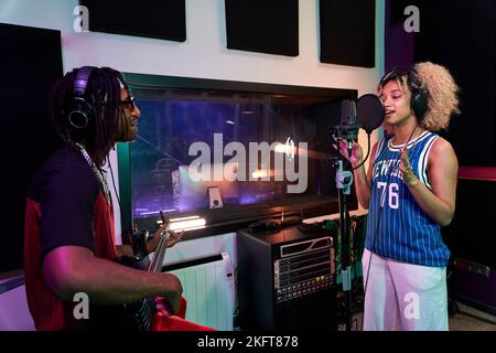 Un musicien afro-américain dans un casque jouant de la guitare tandis qu'un partenaire féminin enregistra une chanson sur le microphone Banque D'Images