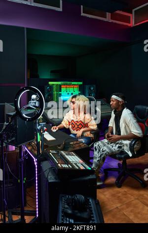 Collègues multiraciaux concentrés assis dans un studio d'enregistrement contemporain avec éclairage au néon lumineux tout en travaillant sur de nouvelles chansons Banque D'Images