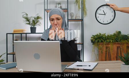 Femme d'affaires programmeur développeur de logiciels travaillant sur ordinateur portable au bureau avec l'inquiétude de vérifier l'heure sur l'horloge, étant en retard, échéance. Jeune fille d'affaires indépendante musulmane concentrée qui s'inquiète d'être en retard Banque D'Images