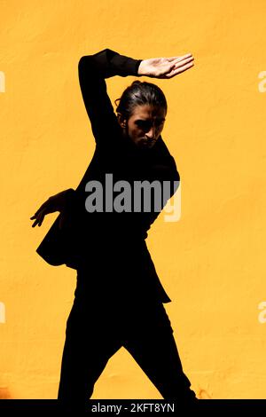 Homme hispanique en costume noir avec des vêtements foncés levant le bras et dansant le flamenco contre le mur jaune Banque D'Images