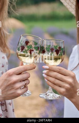 Lunettes à motif. Coupe courte de deux femmes qui applaudissent avec des verres de vin blanc non alcoolisé fait maison tout en se tenant dans un champ de lavande. Banque D'Images