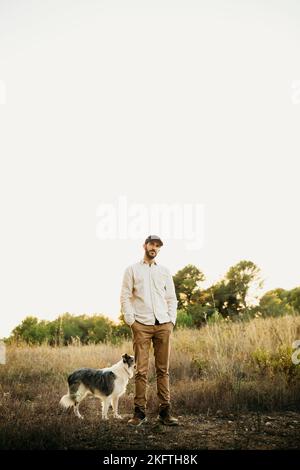 Portrait d'un jeune homme barbu portant une casquette noire, avec son chien, à la campagne Banque D'Images