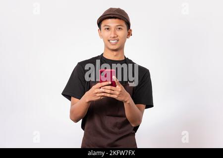 Beau barista asiatique portant un tablier marron et un t-shirt noir isolé sur fond blanc souriant à l'aide d'un smartphone. tenir le téléphone portable pour envoyer des sms Banque D'Images