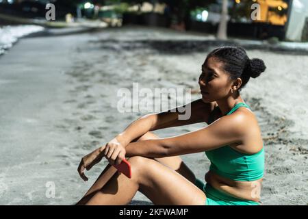 Femme noire dans les vêtements de sport, repose pendant la pause dans l'entraînement de remise en forme sur la plage. Banque D'Images
