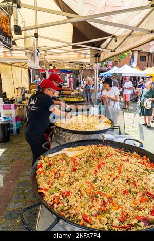 Regardez le long d'un marché en plein air dans un marché français en Angleterre avec une rangée de grands poêles à frire de nourriture en cours de cuisson avec les clients faisant la queue pour acheter. Banque D'Images