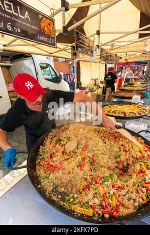 Un panier repas extérieur avec une rangée de grands poêles à frire cuisant divers aliments, avec un homme en premier plan en remuant le premier, plein de paella vapeur. Banque D'Images