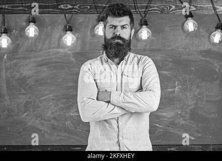 Homme avec la barbe et la moustache sur le visage strict se tenir devant le tableau de surveillance. Hipster barbu en chemise, tableau noir et ampoules sur fond. Strict Banque D'Images