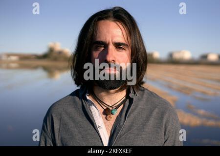 Jeune homme caucasien avec la barbe et les cheveux longs debout regardant la caméra sérieusement à la périphérie de la ville Banque D'Images