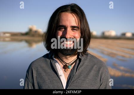 Jeune homme caucasien avec une barbe et de longs cheveux debout souriant aboyant dents à la caméra à la périphérie de la ville Banque D'Images