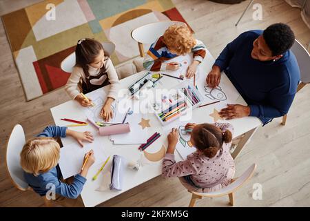 Photo ci-dessus de plusieurs apprenants interculturels de l'école maternelle et de leur enseignant assis devant eux pendant la leçon de dessin Banque D'Images
