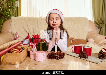 Adorable petite fille en chapeau de père Noël, garde les mains paumes ensemble, fait le souhait, s'assoit à la table avec une bougie et cadeaux de Noël Banque D'Images