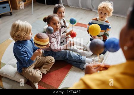 Groupe d'apprenants interculturels sérieux de l'école primaire tenant des modèles de planètes du système solaire tout en étant assis sur le sol devant l'enseignant Banque D'Images