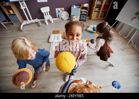 Jolie petite fille gaie avec un modèle de soleil jaune debout parmi ses amis avec des pplanètes et courir autour d'elle tout en jouant au système solaire Banque D'Images