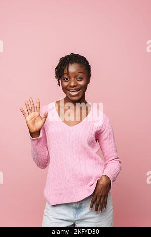 Femme noire excitée avec des dreadlocks agitant la main et souriant à la caméra isolée sur fond rose Banque D'Images