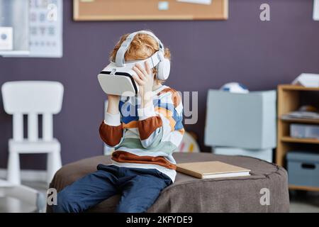 Petit garçon aux cheveux d'épices en pull-over rayé et casque vr jouant au jeu virtuel tout en étant assis sur un pouf rond doux dans une école maternelle Banque D'Images