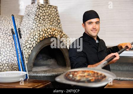 Chef latino en uniforme noir tenant une pelle avec pizza dans le restaurant de cuisine Banque D'Images