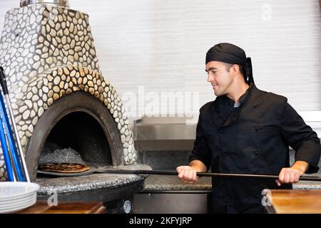 Chef latino en uniforme noir tenant une pelle avec pizza dans le restaurant de cuisine Banque D'Images