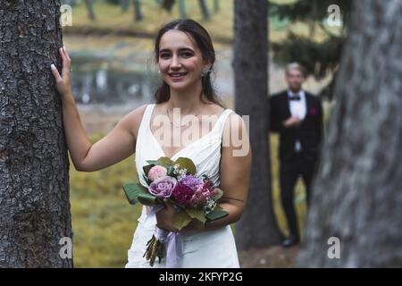 Séance photo d'automne. Photo extérieure moyenne d'une femme turque à la mi-20s, touchant un arbre, souriant et tenant son bouquet de mariée. Silhouette floue d'un élégant marié scandinave en arrière-plan. Photo de haute qualité Banque D'Images