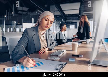 Femme d'affaires musulmane ciblée dans le hijab travaillant avec des documents tout en étant assis dans un bureau moderne Banque D'Images