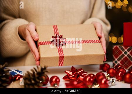 Femme en chandail chaud tenant une boîte cadeau emballage en papier artisanal avec ruban de fête. Cadeau pour les vacances d'hiver. Noël et bonne année Banque D'Images