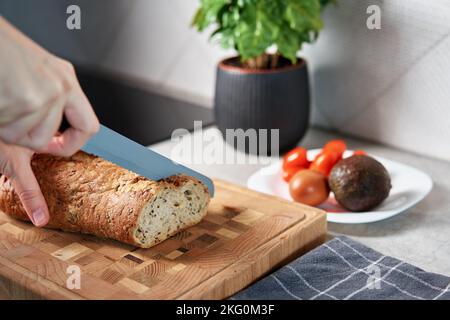 Femme coupant du pain de grain entier avec un grand couteau sur la planche à découper. Tranchez du pain frais et croustillant aux mains dans la cuisine Banque D'Images