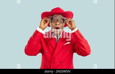 Homme africain en costume rouge et lunettes de fête regardant quelque chose avec l'expression du visage surprise Banque D'Images