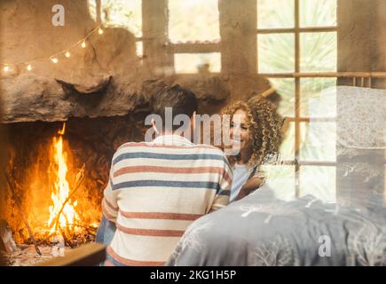 Un couple homme et une femme aiment les loisirs en intérieur assis devant une cheminée chaude pour chauffer la maison. Des jeunes adultes heureux voient à travers un gla Banque D'Images