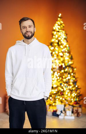 Un jeune homme souriant à capuche se dresse contre le fond d'un arbre de Noël et de cadeaux. Concept de Noël. Arrière-plan orange. Banque D'Images