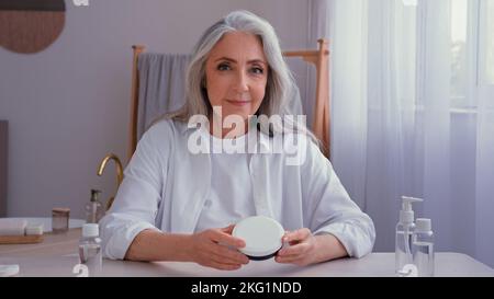 Caucasien vieux senior mature 50s femme avec cheveux gris blogger à l'intérieur à la maison parlant regardant l'appareil photo montrant des produits de beauté crème Banque D'Images