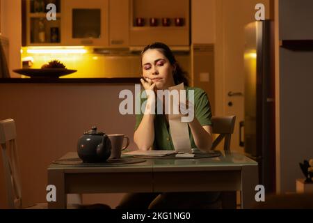 Une jeune femme de race blanche réfléchie est assise à table et vérifie les dépenses à la réception. Comptabilité à domicile. Ambiance de soirée. La femme au foyer est engagée Banque D'Images