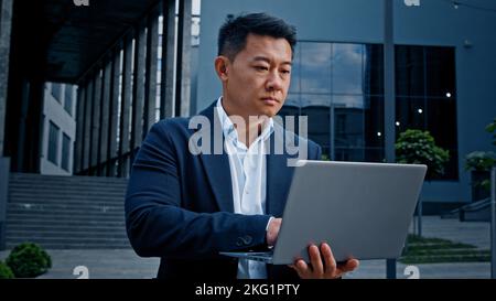 Asiatique 40s homme d'affaires d'âge moyen dactylographiant un ordinateur portable en plein air dans le centre-ville coréen adulte entrepreneur employeur homme travailleur gestionnaire investisseur architecte debout Banque D'Images