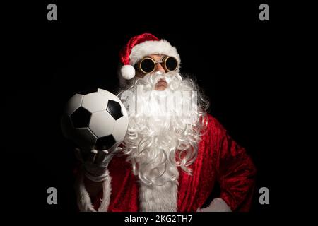Le Père Noël avec des lunettes de soleil ou un soudeur tenant une balle de football de la coupe du monde comme cadeau pour tous les enfants, avec un fond noir où son costume rouge et whi Banque D'Images