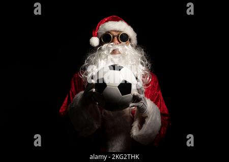 Le Père Noël avec des lunettes de soleil ou un soudeur tenant une balle de football de la coupe du monde comme cadeau pour tous les enfants, avec un fond noir où son costume rouge et whi Banque D'Images