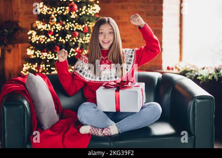 Portrait complet du corps de fille ravie surjoyeuse assis canapé lève les poings reçoivent boîte-cadeau festive à l'intérieur Banque D'Images