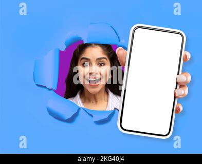 Femme excitée posant dans un mur bleu déchiré, montrant le smartphone Banque D'Images