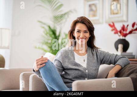 Portrait d'une belle femme aux cheveux longs portant des vêtements décontractés tout en se relaxant dans un fauteuil à la maison. Banque D'Images