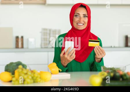 Femme musulmane mûre souriante utilisant un smartphone et une carte de crédit tout en cuisinant dans la cuisine, en commandant des provisions sur Internet Banque D'Images